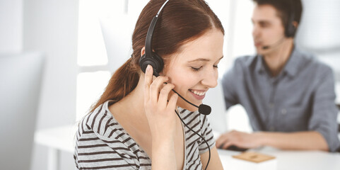 Casual dressed young woman using headset and computer while talking with customers online. Group of operators at work. Call center, business concept