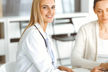 Woman-doctor at work in sunny hospital is happy to consult female patient. Blonde physician checks medical history record and exam results while using clipboard