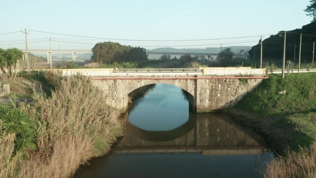 Reflections Through Calm River With Stone Arch Bridge During Daytime At Alcobaca River Near Nazare In Portugal. - Aerial Pullback Shot 