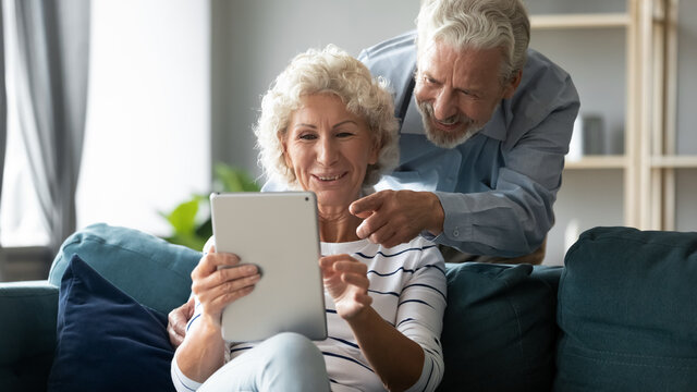 Emotional joyful middle aged senior family couple watching funny photos or videos online on digital computer tablet, having fun web surfing information or enjoying shopping in internet store.