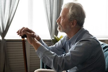 Naklejka na ściany i meble Thoughtful unhappy elderly senior grandfather sitting on cozy sofa with wooden cane in hands, looking in distance. Pensive stressed middle aged disabled old man suffering from loneliness at home.