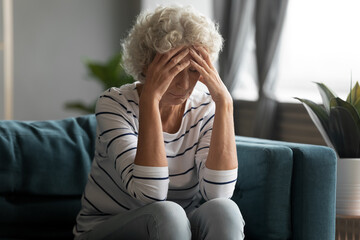 Unhealthy old senior retired woman sitting on sofa, suffering from strong headache. Unhappy elderly...