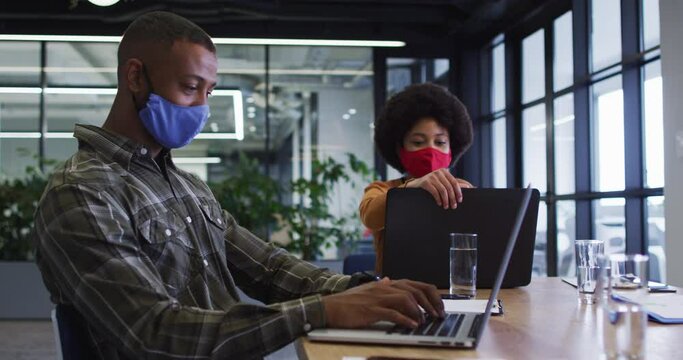 Diverse business people wearing face masks sitting using laptops going through paperwork in office