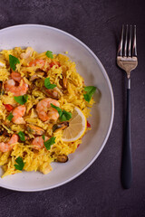 Paella with shrimps, mussels, lemon, herbs in a gray plate next to a fork on a dark gray stone background.