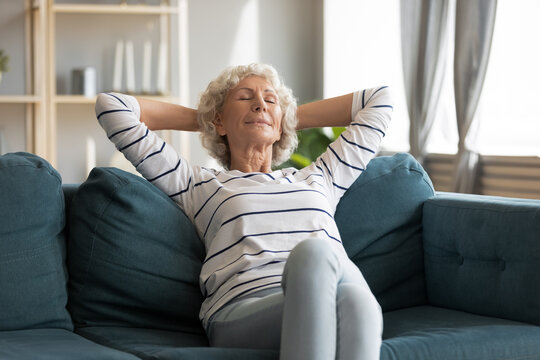 Happy Middle Aged Elderly Hoary Woman Relaxing On Comfortable Couch With Folded Hands Behind Head, Sleeping Or Napping Alone Indoors. Peaceful Calm Retired Grandmother Enjoying Mindful Moment At Home.