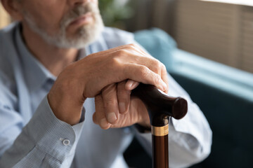 Close up old disabled man holding wrinkled hands on walking cane, feeling loneliness at home....