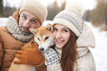 Close up portrait of happy adult couple holding cute dog and looking at camera while enjoying walk outdoors together in winter forest