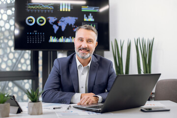 Naklejka premium Front view of mature middle aged confident high-skilled bearded office manager or boss, wearing formal suit, posing on camera in his modern office room, sitting at the desk with laptop pc