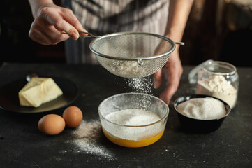 The process of preparing the dough. Ingredients for the dough on a dark background. In women's hands a sieve from which flour is poured.
