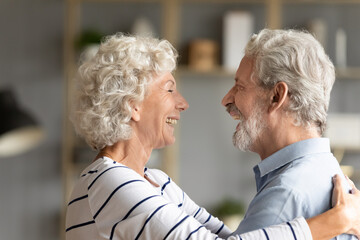 Happy hoary middle aged woman cuddling affectionate senior husband, missing glad to see after long separation. Smiling old mature family couple showing tender sweet feelings to each other at home.