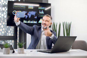 Handsome happy bearded middle aged businessman making selfie photo on the smartphone, while sitting at the table with laptop in modern office. Big wall digital screen on the background