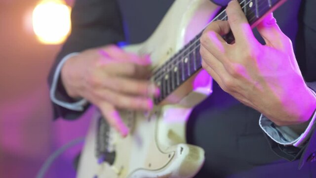 Close up of a professional musician in a black suit playing fast chords on an electric stratocaster guitar during a live session on stage with warm studio lights in the blurred background. Shot in 4K.