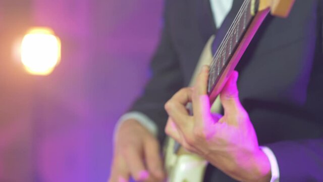 Close up of a professional musician in a black suit playing chords on an electric stratocaster guitar during a live session on stage with warm studio lights in the blurred background. Shot in 4K.