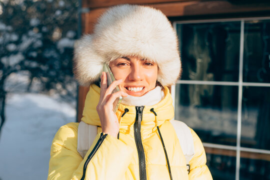 Young beautiful happy woman outdoors on a sunny day with a phone in warm clothes and a winter russian siberian hat