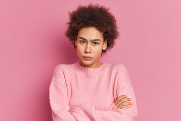 Fototapeta na wymiar Portrait of serious angry African American woman stands with crossed hands looks irritated at camera waits for your explantions wears casual sweater isolated over pink background. Negative emotions