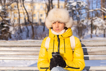 A woman in winter in warm clothes in a snow-covered park on a sunny day sits on a bench and is...