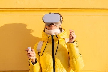 Portrait of a woman wearing virtual reality glasses on the street against a yellow wall in bright sunlight in winter in warm clothes