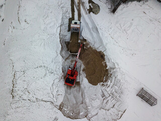 An excavator loads sand into a truck. Snowy day, blizzard. Aerial drone top view.