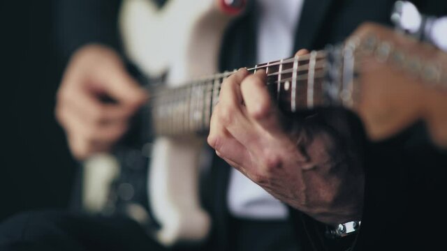 Close up of a professional musician in a black suit playing chords on a white electric stratocaster guitar during a live session with a dark background. Shot in 4K.