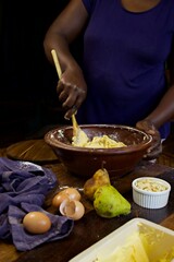 a black person stiring cake batter in a wooden bowl at a kitchen table.  A blue cloth, eggshells and butter sits in the foreground