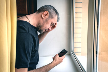 Portrait of a man using a smartphone in front of a window. Upset man reading morning news on smartphone