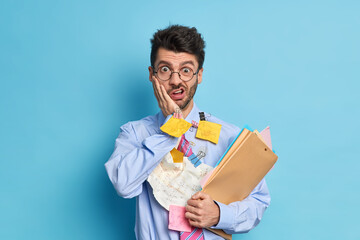 Studio shot of male employee stares surprised at camera busy doing paper work creats business plan holds folders with papers keeps hand on cheek wears formal clothing isolated over blue background