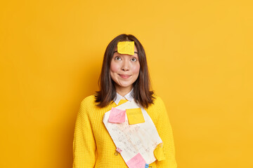 Pretty Asian schoolgirl has sticky notes and papers with written formulas stuck on clothes looks above crams for exam wears casual sweater isolated over yellow studio background. Studying concept