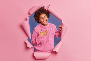 Happy sincere young African American woman giggles positively feels very glad keeps hand on chest wears knitted sweater breaks through paper hole with ripped sides. Positive emotions concept