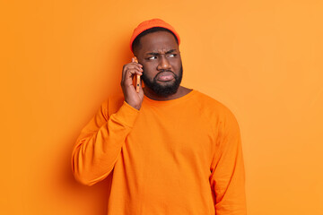 Serious bearded Afro American man has phone conversation talks via smartphone and looks thoughtfully aside wears hat and casual sweater poses against orange background. Communication concept