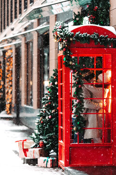 Stylish romantic couple kissing, hugging, having fun in winter before Christmas, standing in vintage red telephone booth during snowfall. New Year Eve concept. 