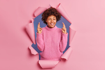 Positive dark skinned young woman crosses fingers winks eye makes wish smiles broadly dressed in knitted sweater breaks through paper background hopes dreams come true believes in good luck.