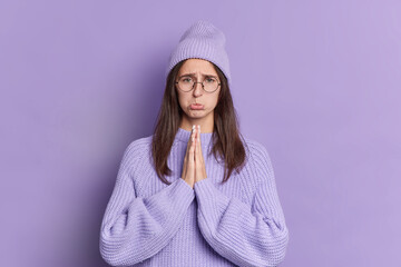 Sad brunette woman looks with pleading expression at camera keeps palms together asks for apology...
