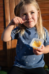 Niña rubia comiéndose un flan con su casa de madera al fondo en un atardecer de verano