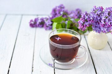 Delicate setting of the morning tea table with lilac flowers, a transparent cup of tea and a saucer and a white vase on a white wooden board. Copy space. Spring breakfast concept