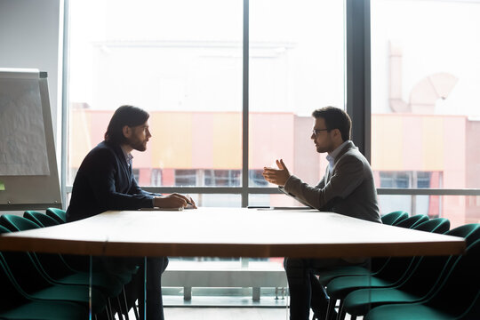 Focused Confident Young Businessman Making Offer To Concentrated Male Partner At Negotiations Meeting In Modern Office. Two Successful Entrepreneurs Discussing Project Details, Briefing Indoors.