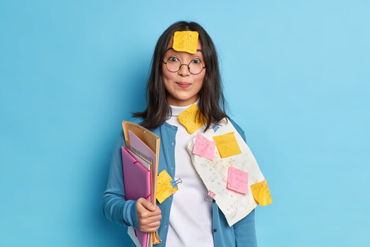 Pretty schoolgirl prepares for math test crams material has sticker on forehead not to forget necessary information busy studying isolated over blue background. Asian woman with paper notes.