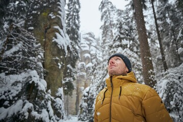 Fototapeta na wymiar Portrait of young man in winter nature. Tourist in warm clothing against rocks in the middle forest during snowing. Adrspach, Czech Republic