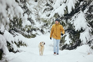Front view of young man with dog in winter nature. Pet owner with his labrador retriever are walking in snow covered forest. 