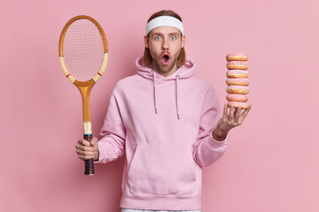 Bearded shocked hipster wears sport outfit holds tennis racket and pile of sweet doughnuts gazes...