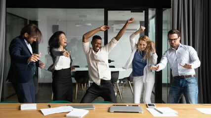 Crazy emotional young african american man having fun with happy diverse mixed race colleagues, dancing together in modern office, celebrating corporate success or profitable goal deal achievement. - Powered by Adobe