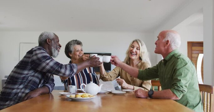 Two diverse senior couples sitting by a table drinking tea together making a toast at home