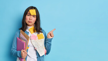Horizontal shot of unhappy brunette Asian woman holds folders with papers gives recommendations how to prepare for exams has sticker on forehead indicates at copy space over blue background.