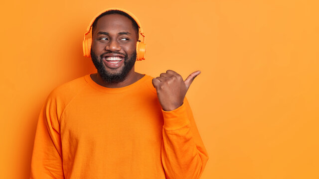 Studio Shot Of Joyful Bearded Black Man Listens Audio Track Via Wireless Headphones Wears Bright Orange Long Sleeved Jumper In One Tone With Background Points Thumb Away Advertises Copy Space