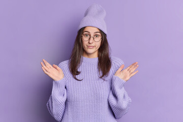 Photo of clueless millennial girl with long dark hair spreads palms and poses unaware indoor has no idea wears round transparent glasses knitted hat sweater isolated over purple background. Monochrome