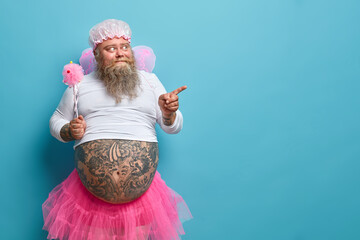 Surprised bearded man with fat tattoed belly wears fairy costume holds magic wand indicates away...