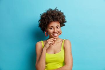 Portrait of pleasant looking curly woman keeps hand under chin smiles gently feels satisfied...