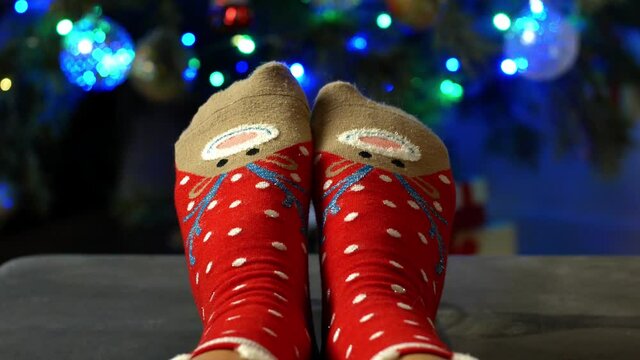 Woman legs in cozy Christmas socks with a deer on the background of a decorated Christmas tree. Close-up of Christmas socks and tree for Christmas 4K video.