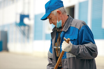 This is an elderly janitor with a broom in a medical mask on the street sweeping the territory. An...
