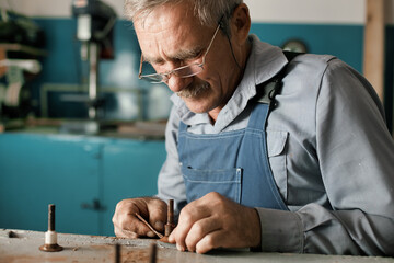 An old master with glasses works in the workshop. A white elderly man in work clothes sits at a workbench and carefully examines a metal part. Artistic photo with tinted effect