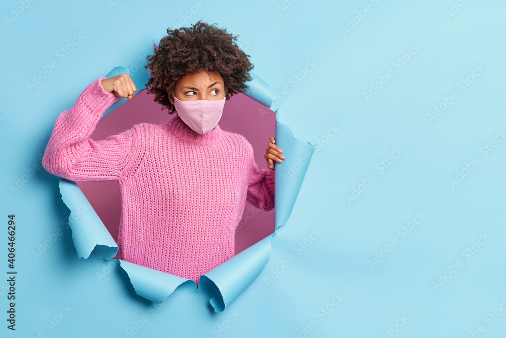 Wall mural Thoughtful dark skinned young woman raises arm shows muscles wears protective face mask during coronavirus pandemic dressed in knitted sweater poses through ripped hole looks pensively aside